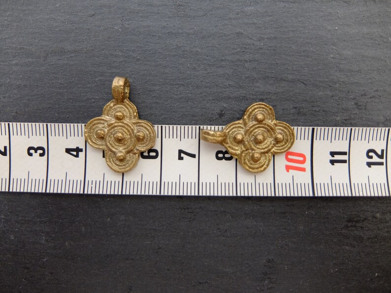 May include: Two antique-style, gold-colored pendants with a four-petal flower design. Each pendant features concentric circles and small spheres. The pendants are placed on a measuring tape, with one pendant measuring approximately 1.5 inches in length.