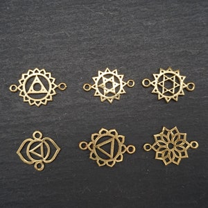 Könnte beinhalten: Sechs goldfarbene Metall-Anhänger mit verschiedenen Chakra-Symbolen. Die Anhänger sind so konzipiert, dass sie als Verbindungselemente für die Schmuckherstellung verwendet werden können.