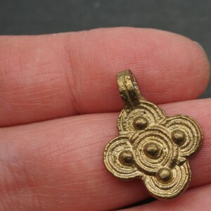 May include: A small, antique-style brass pendant with a four-lobed design. Each lobe features concentric circles and a central raised dot. The pendant has a loop at the top for hanging. The overall design is symmetrical and detailed.