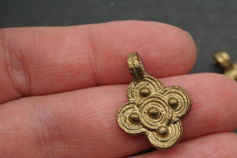 May include: A small, antique-style brass pendant with a four-lobed design. Each lobe features concentric circles and a central raised dot. The pendant has a loop at the top for hanging. The overall design is symmetrical and detailed.