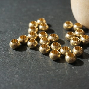 Könnte beinhalten: Eine Gruppe von 20 goldfarbenen Metallperlen, die auf einer dunkelgrauen Oberfläche verstreut sind. Die Perlen sind rund und haben ein kleines Loch in der Mitte.