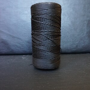 May include: A spool of black waxed nylon cord, tightly wound. The cord is cylindrical and appears to be new. The background is a dark gray, textured surface. This cord is suitable for various crafting and jewelry-making projects.