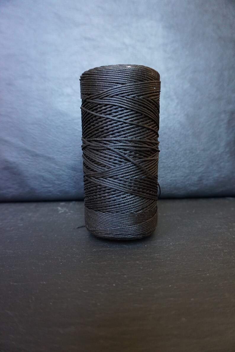 May include: A spool of black waxed nylon cord, tightly wound. The cord is cylindrical and appears to be new. The background is a dark gray, textured surface. This cord is suitable for various crafting and jewelry-making projects.