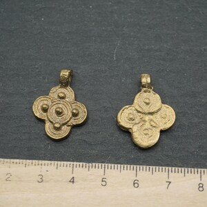 May include: Two antique gold-colored pendants with a four-petal floral design. Each pendant has a loop at the top for hanging. The pendants are displayed on a dark gray surface next to a ruler.