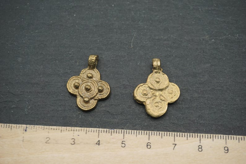 May include: Two antique gold-colored pendants with a four-petal floral design. Each pendant has a loop at the top for hanging. The pendants are displayed on a dark gray surface next to a ruler.