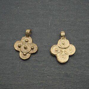 May include: Two antique gold-colored pendants with a four-petal floral design. Each pendant has a loop at the top for hanging. The pendants are displayed on a dark gray, textured surface.