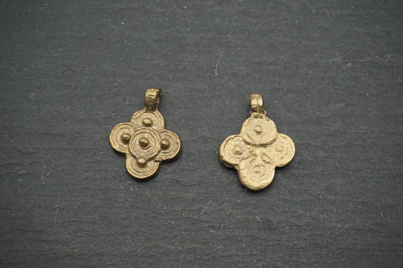 May include: Two antique gold-colored pendants with a four-petal floral design. Each pendant has a loop at the top for hanging. The pendants are displayed on a dark gray, textured surface.