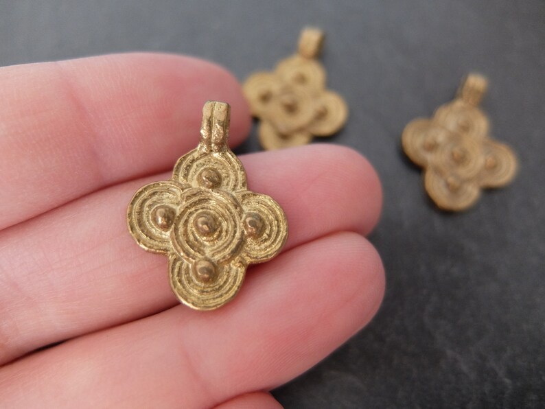 May include: Close-up of several small, gold-colored pendants. The pendants have a four-petal flower design with concentric circles and small, raised dots. The pendants are likely made of metal and are suitable for jewelry making.