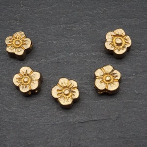 Könnte beinhalten: Fünf goldfarbene Metallperlen in Blumenform. Die Perlen haben eine erhabene Mitte und fünf Blütenblätter.