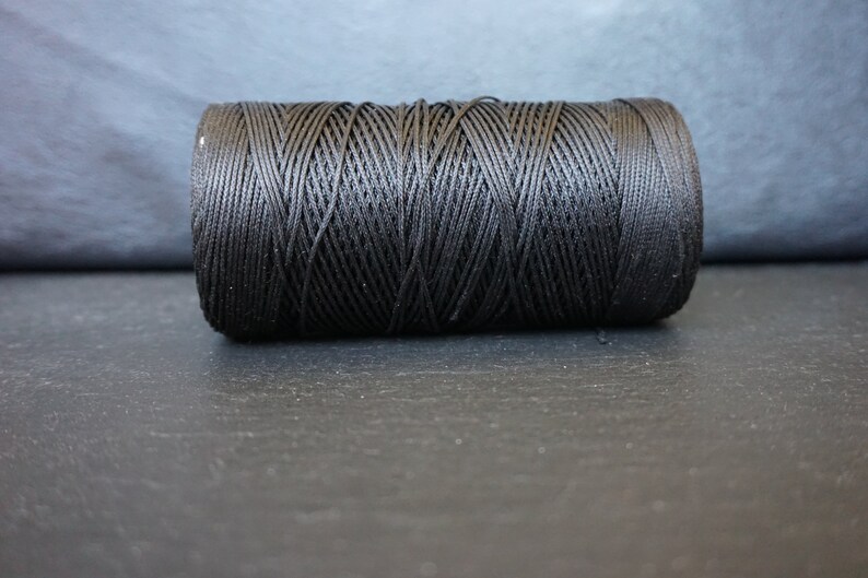 May include: A spool of black thread, tightly wound and cylindrical in shape. The thread is a deep, solid black, and the texture of the wound thread is clearly visible. The spool is resting on a dark surface.