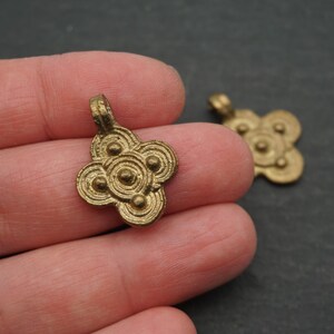 May include: Two small, antique-style brass pendants. Each pendant has a four-petal floral design with circular details. The pendants are a warm gold color and have a loop at the top for hanging.