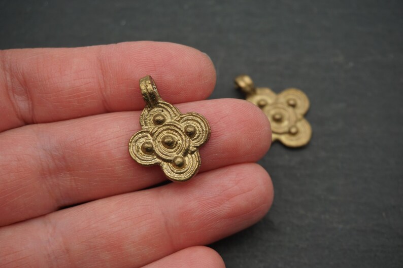 May include: Two small, antique-style brass pendants. Each pendant has a four-petal floral design with circular details. The pendants are a warm gold color and have a loop at the top for hanging.
