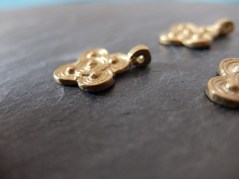 May include: Close-up of several small, gold-colored cloud-shaped pendants. Each pendant has a loop for hanging and a detailed, swirling design. The pendants are on a dark gray surface.
