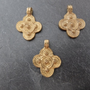 May include: Three gold-colored pendants with a four-petal flower design. Each pendant features a loop at the top for hanging and a series of concentric circles and small spheres within the petals. The pendants are displayed on a dark gray surface.