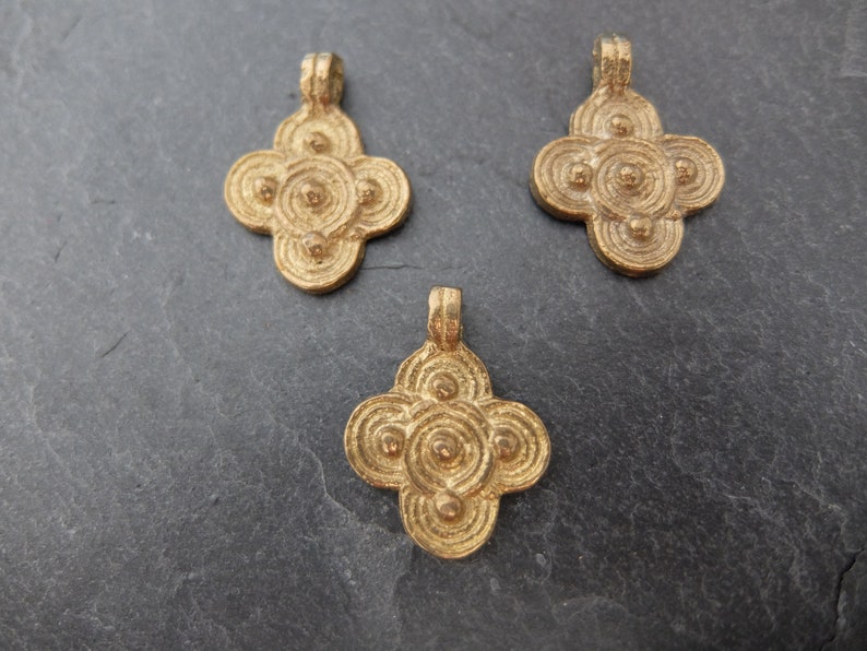 May include: Three gold-colored pendants with a four-petal flower design. Each pendant features a loop at the top for hanging and a series of concentric circles and small spheres within the petals. The pendants are displayed on a dark gray surface.
