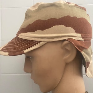 以下が含まれることがあります： 茶色とベージュの迷彩柄の軍用スタイルの帽子。耳と首を覆うようにフラップを下ろすことができます。