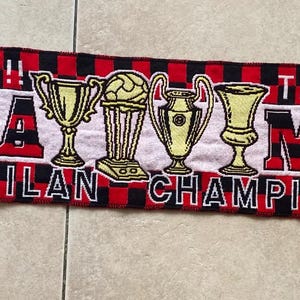 May include: A red and black checkered knit scarf featuring the text THE BEST!! FORZA MILAN THE WINNERS!! MILAN CHAMPION. The design includes four gold trophy illustrations and two yellow star graphics with soccer balls on the ends with fringe.