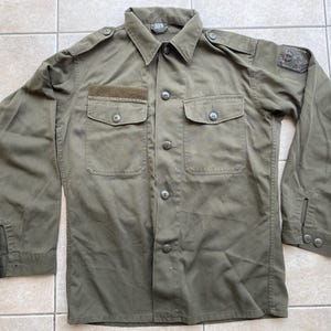 Op de afbeelding: Olijfgroen shirt met lange mouwen, twee borstzakken en knoopsluitingen. Het shirt heeft een kraag, epauletten en een kleine patch op de linkermouw. De stof lijkt een duurzaam, mogelijk militair materiaal te zijn.