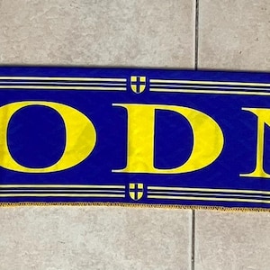 May include: A blue scarf with yellow fringe and the word "MODNA" in large yellow letters. A yellow illustration of a vehicle is on the left side. The scarf has a repeating pattern of small white crosses.