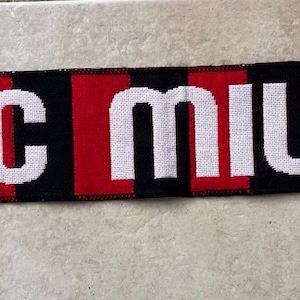 May include: A knitted scarf featuring vertical red and black stripes with the text AC MILAN in white letters across the centre. The scarf includes the AC Milan team crest on both ends and is finished with red, black, and white fringe at each side.