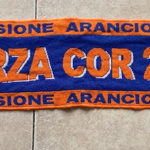 May include: A rectangular knit scarf featuring horizontal orange and blue stripes with fringed ends. The center displays the orange text FORZA COR 2005, flanked by two circular emblems depicting the Colosseum. The text PASSIONE ARANCIO BLU appears above and below the main lettering.