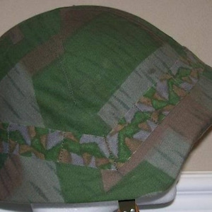 Könnte beinhalten: Ein grüner, brauner und weißer Tarnhelm mit Stoffbezug. Der Helm hat einen breiten Rand und einen Riemen.