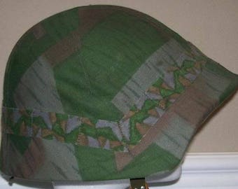 Può includere: Un elmetto mimetico verde, marrone e bianco con una copertura in tessuto. L'elmetto ha un'ampia visiera e un cinturino.