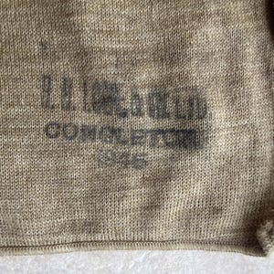 Puede incluir: Primer plano de una tela de punto beige con texto negro descolorido. El texto parece leer "L.H. LLOYD CONSOLIDATED" y otro texto ilegible. La tela tiene un aspecto texturizado.