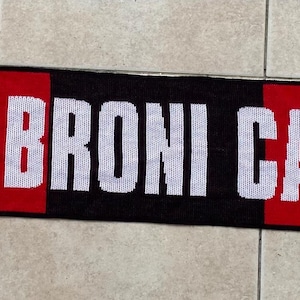 May include: A black, red, and white scarf with the text "ASD BRONI CALCIO" in white. The scarf has a red and black design with a circular logo on each end featuring a soccer player.