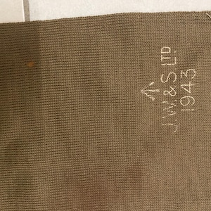 May include: A close-up shot of a brown knitted fabric. The fabric has the embroidered text "J.W.& S LTD 1943" in white. The fabric has a textured appearance and a small stain. The item appears to be a piece of vintage clothing or textile.