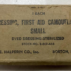 Puede incluir: Una caja rectangular de color marrón claro con la etiqueta "DRESSING, FIRST AID CAMOUFLAGED SMALL". La caja también dice "DYED DRESSING-STERILIZED" y "A. E. HALPERIN CO., Inc., BOSTON, MASS." La caja es vintage y muestra desgaste.