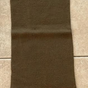 Peut inclure: Une écharpe tricotée de couleur vert olive avec une texture côtelée verticale. Le coin inférieur présente un marquage blanc imprimé avec un symbole de flèche pointant vers le haut au-dessus du texte J.W.& S. Ltd et l'année 1940 en caractères clairs.