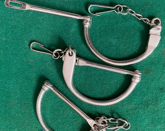 Puede incluir: Tres pares de esposas de metal plateado con una cadena que conecta dos de ellas. Las esposas están abiertas y sobre una superficie verde.