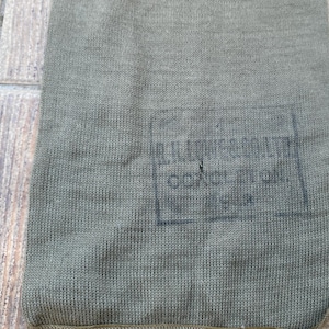 May include: Close-up of an olive green knitted fabric with a rectangular printed design. The design appears to be text within a rectangular border. The fabric has a textured appearance.