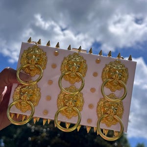 May include: A light pink clutch with gold lion head details and gold ring handles. The clutch has gold spikes along the top edge and gold dot accents. The background shows a blue sky with clouds.