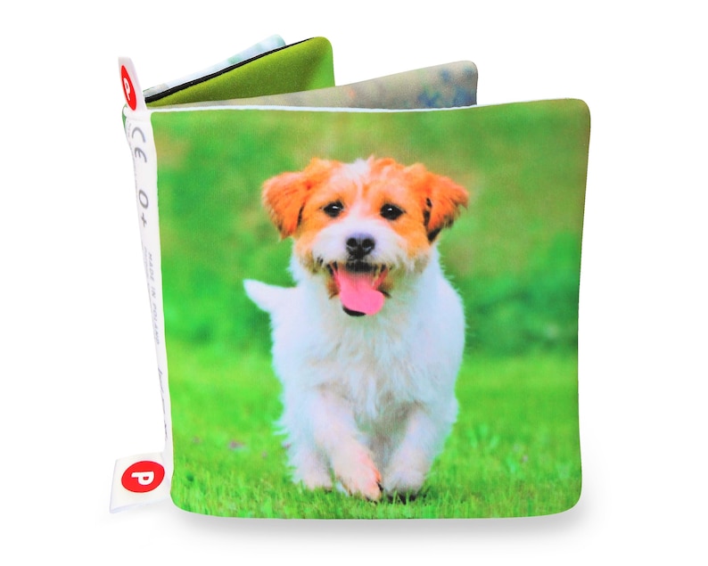 May include: A soft, colorful book for babies and toddlers featuring a white and brown dog running on green grass. The book has a CE 0+ label and a P label.
