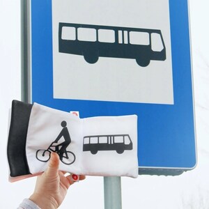 May include: Two white fabric pillows with black designs. One pillow has a black silhouette of a bus, the other has a black silhouette of a person riding a bicycle. The pillows are in front of a blue and white sign with a black silhouette of a bus.