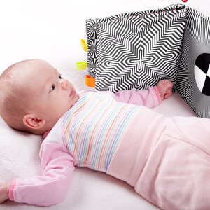 Puede incluir: Un beb&eacute; acostado en una manta blanca mirando un libro de beb&eacute; con patrones en blanco y negro con etiquetas de colores.
