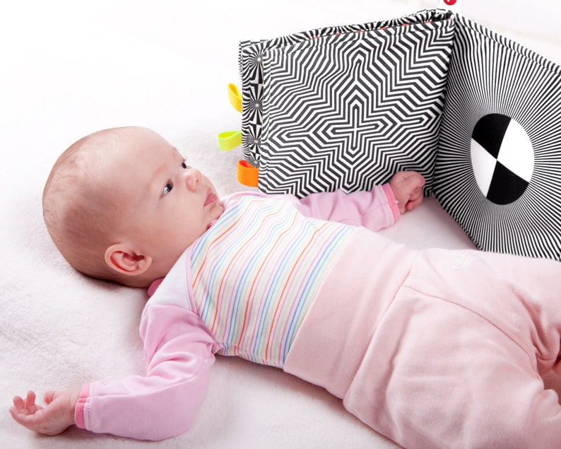 Puede incluir: Un beb&eacute; acostado en una manta blanca mirando un libro de beb&eacute; con patrones en blanco y negro con etiquetas de colores.