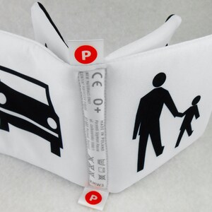 May include: A white fabric cube with black illustrations of a car, a person walking, and a child walking. The cube has a red label with the letter "P" on each side. The label also includes the text "CE 0+", "MADE IN POLAND", "Parents Szymon Face", and washing instructions.