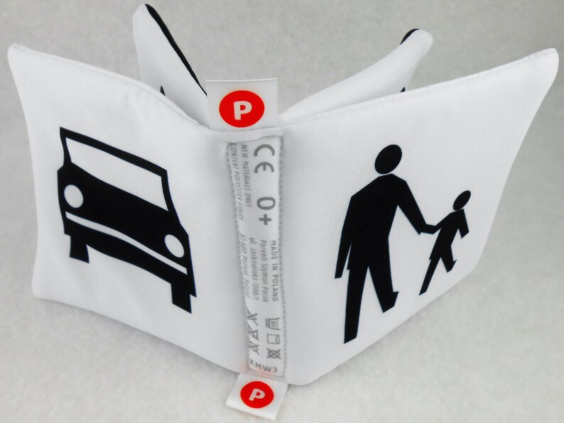 May include: A white fabric cube with black illustrations of a car, a person walking, and a child walking. The cube has a red label with the letter "P" on each side. The label also includes the text "CE 0+", "MADE IN POLAND", "Parents Szymon Face", and washing instructions.