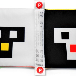 May include: A black and white fabric book with a yellow, green, and pink trim. The book features a pixelated face with black eyes, a yellow mouth, and a red mouth on the opposite page. The book has a CE 0+ label and a "Made in Poland" label.