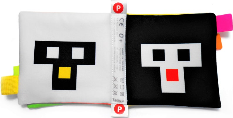 May include: A black and white fabric book with a yellow, green, and pink trim. The book features a pixelated face with black eyes, a yellow mouth, and a red mouth on the opposite page. The book has a CE 0+ label and a "Made in Poland" label.