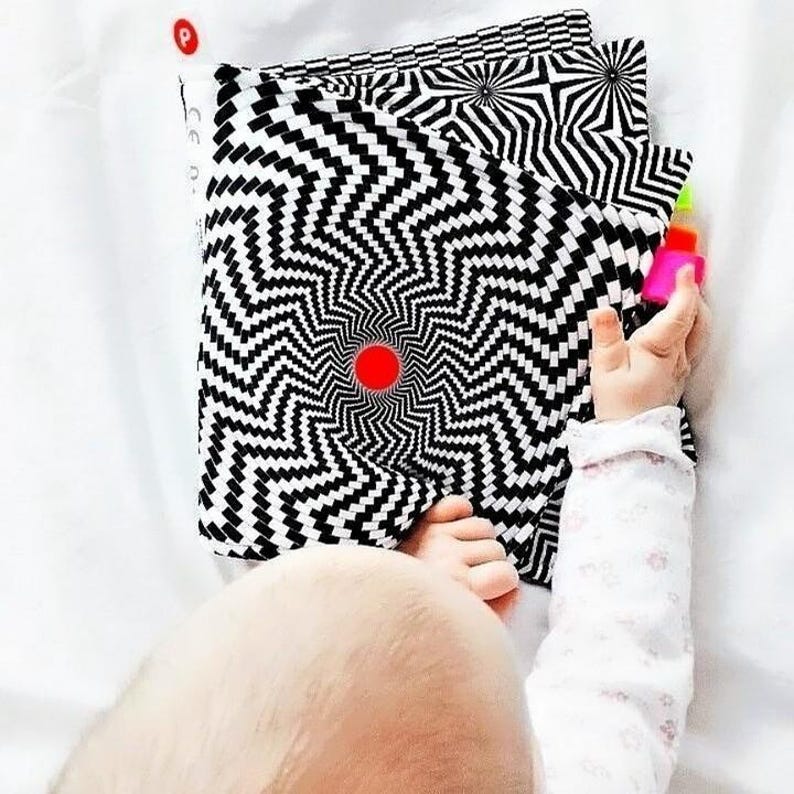 Puede incluir: Un libro con un patr&oacute;n en blanco y negro con un dise&ntilde;o en espiral. Un beb&eacute; est&aacute; sosteniendo el libro y mirando el centro del libro.