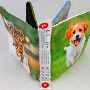 May include: A white fabric book with a cat and a dog on the front covers. The book has a white label with the text "CE 0+" and "MADE IN POLAND".