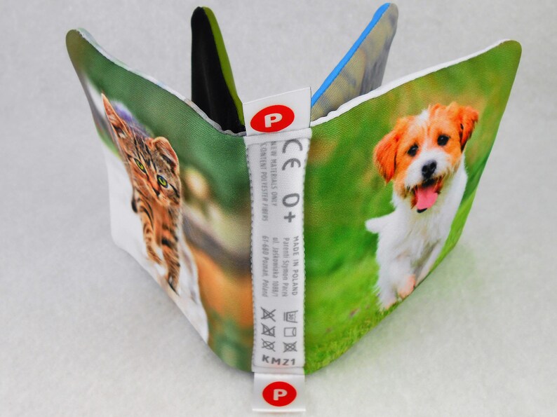 May include: A white fabric book with a cat and a dog on the front covers. The book has a white label with the text "CE 0+" and "MADE IN POLAND".