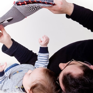 Puede incluir: Un padre y un hijo est&aacute;n tumbados sobre una superficie blanca, leyendo un libro en blanco y negro con acentos rojos y negros. El ni&ntilde;o lleva una camisa a rayas azul y blanca y el padre lleva una camisa negra y gafas.