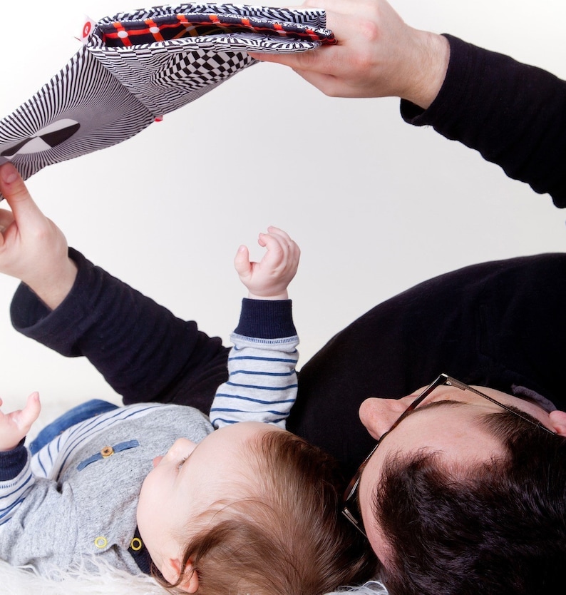 Puede incluir: Un padre y un hijo est&aacute;n tumbados sobre una superficie blanca, leyendo un libro en blanco y negro con acentos rojos y negros. El ni&ntilde;o lleva una camisa a rayas azul y blanca y el padre lleva una camisa negra y gafas.