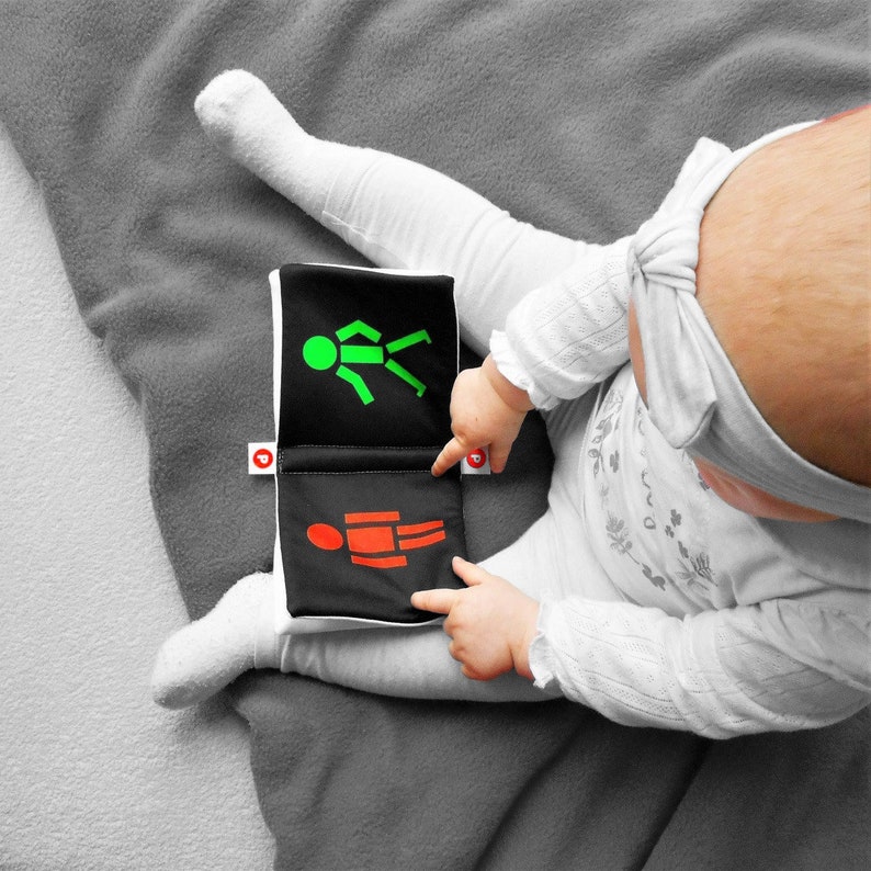 May include: A black and white fabric book with a green and red stick figure on each page. The book is open to the page with the red stick figure. A baby is holding the book and pointing at the red stick figure.
