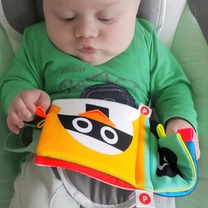 May include: A colorful, multi-textured baby toy with a black and white penguin face. The toy has a yellow, orange, red, green, and blue design. The toy is being held by a baby wearing a green shirt.
